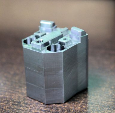 Pieza de plástico gris con detalles y cavidades, posiblemente un componente industrial.